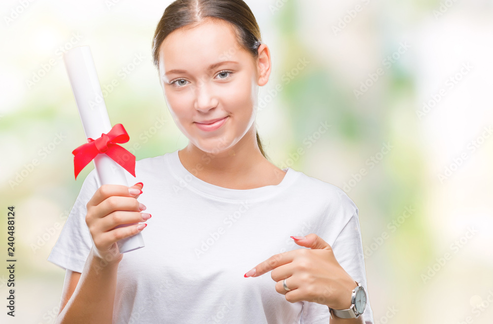 Young caucasian woman holding degree over isolated background with surprise face pointing finger to himself Young caucasian woman holding degree over isolated background with surprise face pointing finger to himself