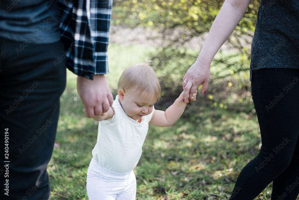 One year old taking first steps, holding both parents hands on a sunny spring day in a park One year old taking first steps, holding both parents hands on a sunny spring day in a park