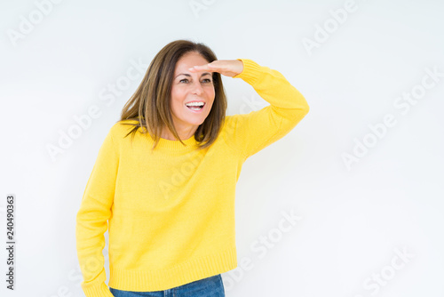 Beautiful middle age woman wearing yellow sweater over isolated background very happy and smiling looking far away with hand over head. Searching concept.
