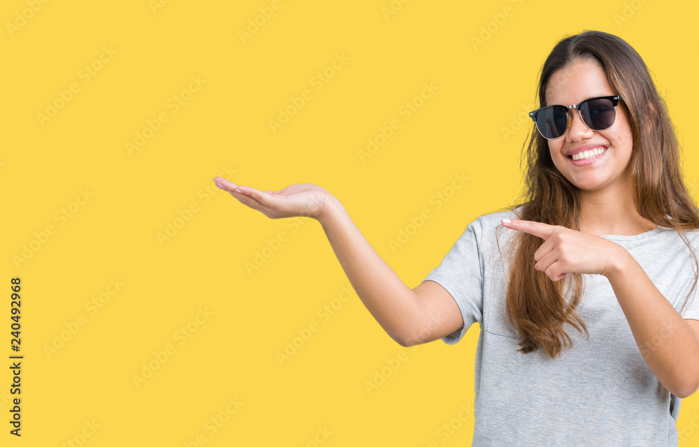 Young beautiful brunette woman wearing sunglasses over isolated background amazed and smiling to the camera while presenting with hand and pointing with finger. Young beautiful brunette woman wearing sunglasses over isolated background amazed and smiling to the camera while presenting with hand and pointing with finger.