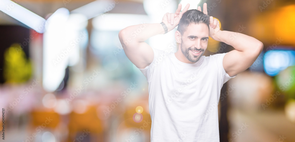 Handsome man wearing white t-shirt over night outdoors background Posing funny and crazy with fingers on head as bunny ears, smiling cheerful Handsome man wearing white t-shirt over night outdoors background Posing funny and crazy with fingers on head as bunny ears, smiling cheerful