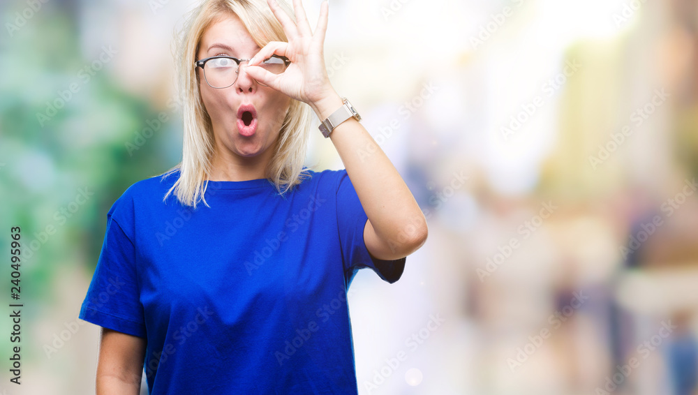 Young beautiful blonde woman wearing glasses over isolated background doing ok gesture shocked with surprised face, eye looking through fingers. Unbelieving expression.
