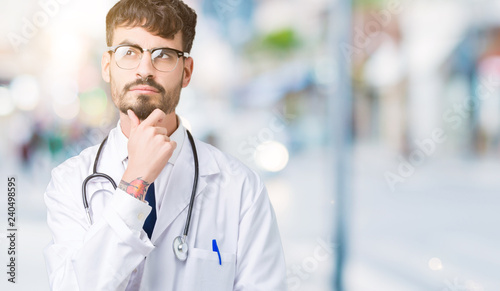 Young doctor man wearing hospital coat over isolated background with hand on chin thinking about question, pensive expression. Smiling with thoughtful face. Doubt concept.