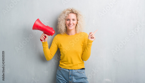 Young blonde woman over grunge grey background holding megaphone very happy pointing with hand and finger to the side