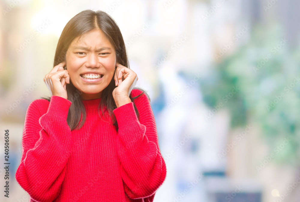 Young asian woman wearing winter sweater over isolated background covering ears with fingers with annoyed expression for the noise of loud music. Deaf concept.