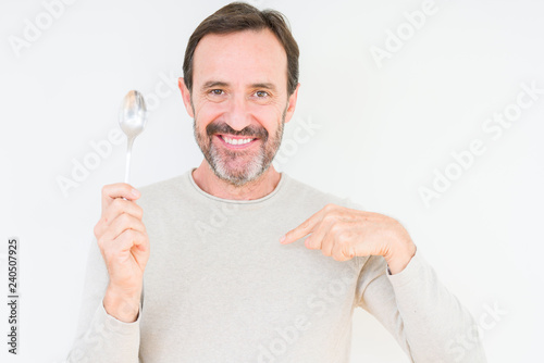 Senior man holding silver spoon over isolated background with surprise face pointing finger to himself