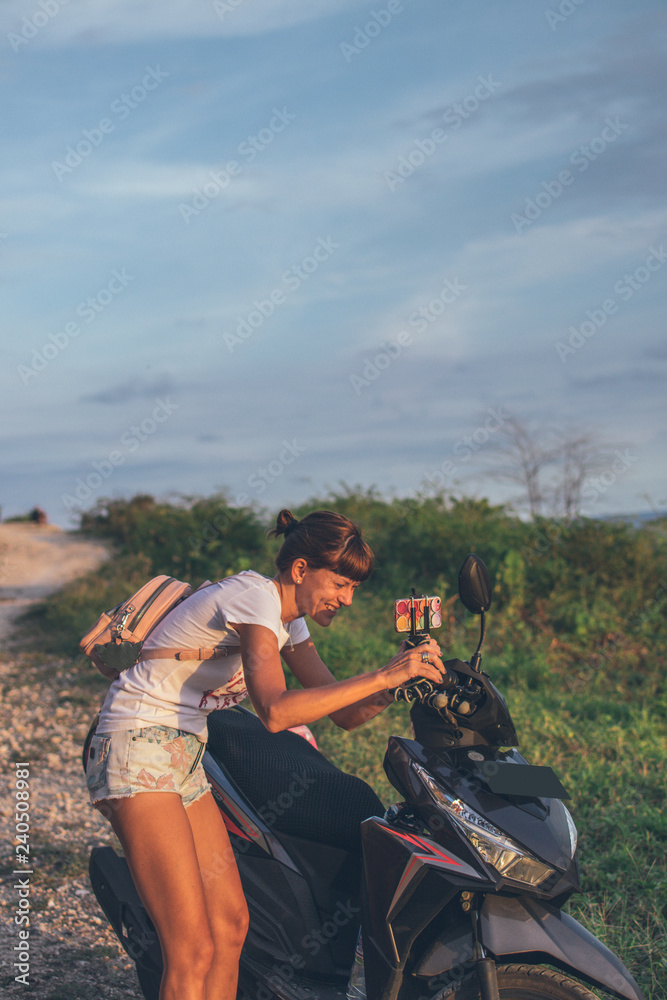 Young woman taking photo of sunset on motorbike. Bali island. Indonesia.