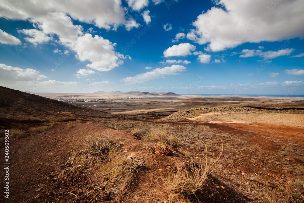 Amazing vulcanic landscape with mountains, ocean and small village Lajares on horizon. Sky is blue with clouds, gravel is red and brown. Fuerteventura Island, Canary Island, Spain, Europe. Amazing vulcanic landscape with mountains, ocean and small village Lajares on horizon. Sky is blue with clouds, gravel is red and brown. Fuerteventura Island, Canary Island, Spain, Europe.