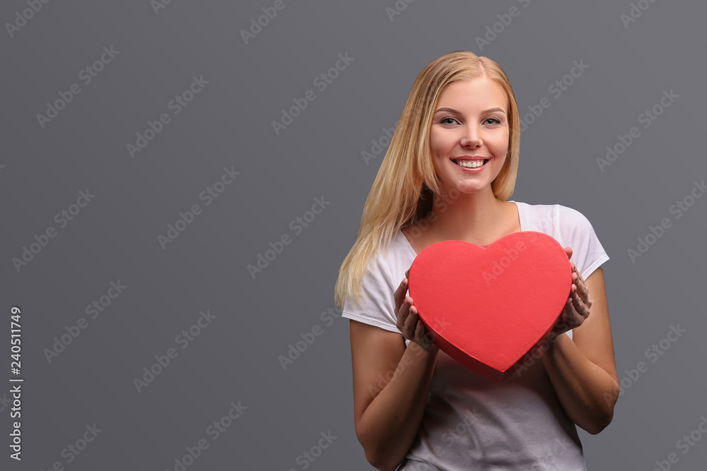 Young blonde girl with gift in hand, red in the shape of a heart. Emotions of joy and surprise on the face. Isolate on a gray background. Young blonde girl with gift in hand, red in the shape of a heart. Emotions of joy and surprise on the face. Isolate on a gray background.