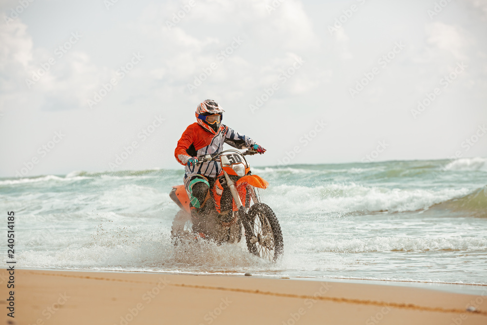 Obraz premium Motorcyclist in a protective suit rides a motorcycle on the sea, splashes fly from under the wheels.