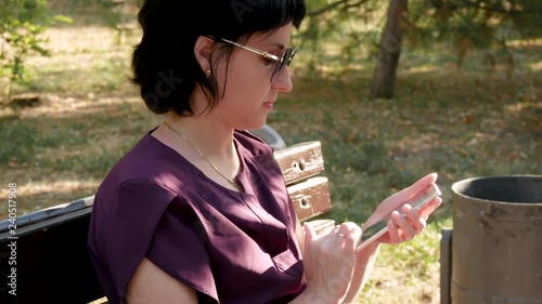 A brunette girl with sunglasses sits on a bench in a summer park and looks at the screen of her phone and touches the sensor eran with her fingers. photo