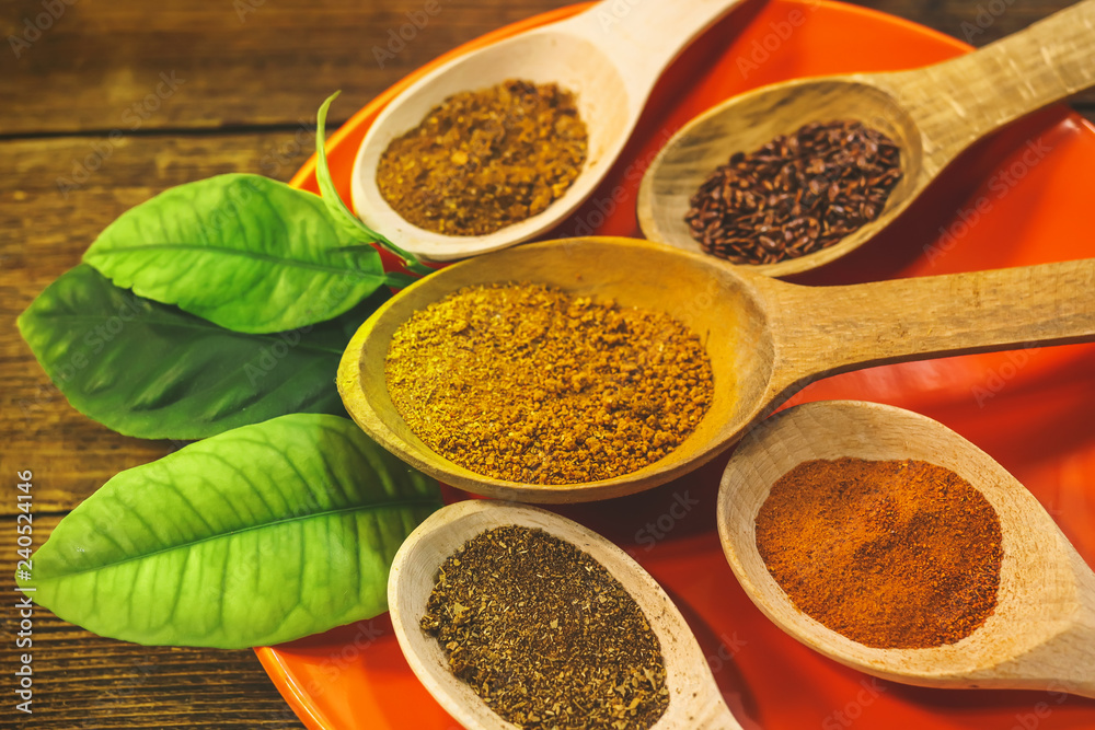 various spices in wooden spoons on an orange plate with lemongrass leaves. kari, chilli peppers, dry spices, flax seeds, pumpkin seeds. wooden table. various spices in wooden spoons on an orange plate with lemongrass leaves. kari, chilli peppers, dry spices, flax seeds, pumpkin seeds. wooden table.