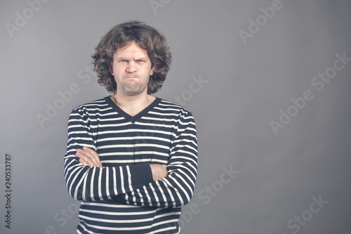 Young man with dark brown hair wears black and white striped casual tshirt looks angry, lips pursed frowning brows, isolated over grey background. Keeps arms crossed and looks at camera. Disagreement photo