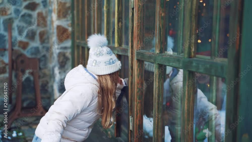 Cheerful little girl with long hair and in warm clothes is peeping through the window from the street behind the elves in the winter against the background of the building. Good New Year spirit Cheerful little girl with long hair and in warm clothes is peeping through the window from the street behind the elves in the winter against the background of the building. Good New Year spirit