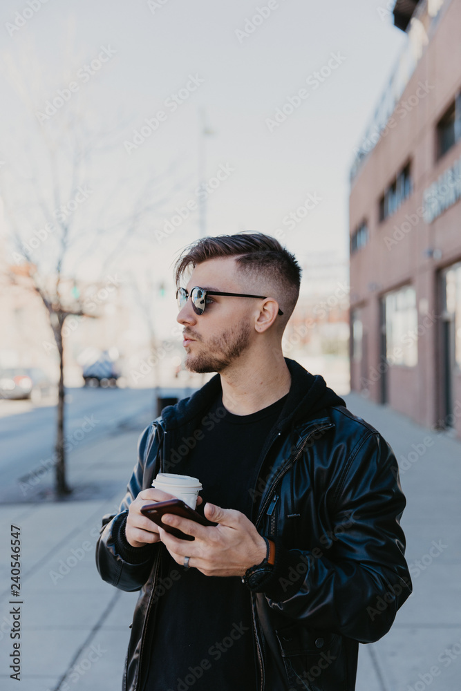 Fototapeta premium Young Happy Millennial Male Business Man Entrepreneur in Urban Downtown Metropolis on His Cell Phone Texting and Taking Business Phone Calls on Walking Commute While Sipping Coffee Beverage To Go