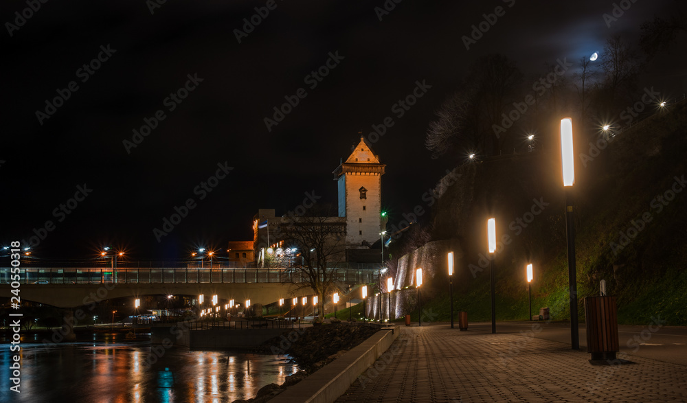 Night view of Narva castle with the tower of High Herman, Narva, Estonia. The castle is illuminated. In the foreground is the city promenade. Opposite Narva is the Russian city of Ivangorod
