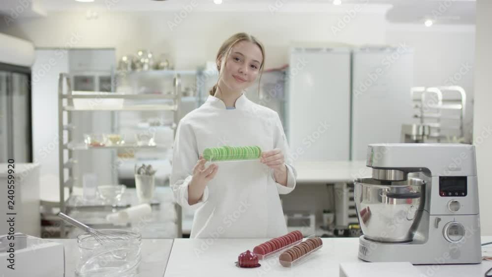 Portrait of a happy young trainee chef or worker in a commercial kitchen. Woman is posing in the kitchen with macarons Portrait of a happy young trainee chef or worker in a commercial kitchen. Woman is posing in the kitchen with macarons