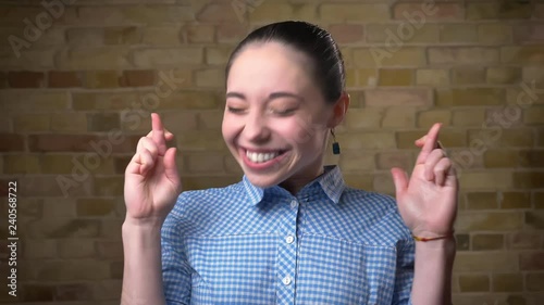 Close-up portrait of caucasian brunette woman with ponytail with crossed fingers gesture showing hope on bricken wall background. photo