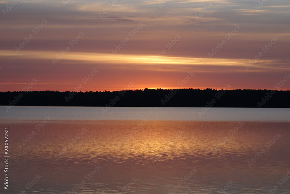 Sunset on the lake. On the horizon the forest. The sun had already fallen over the edge of the forest. The sky is from grey to yellow and red. The forest is dark, the water is also gray and yellow-red