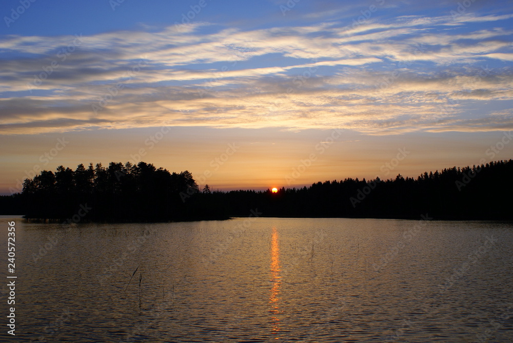 Sunrise on the lake. On the horizon a dark forest. The sun appeared over the tops of the trees and painted the sky pink. Blue sky. The water in the lake is dark blue. In the water trail of the sun.