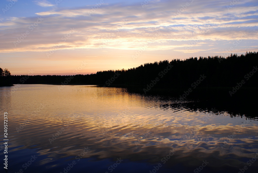 Obraz premium Early morning on the lake. On the horizon a dark forest. The sun has not risen yet, but has painted the clouds pink. Blue sky. The water in the lake is blue, the water reflects pink clouds.
