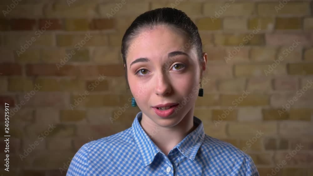 Close-up portrait of caucasian brunette woman with ponytail approvingly nodding into camera on bricken wall background. Close-up portrait of caucasian brunette woman with ponytail approvingly nodding into camera on bricken wall background.