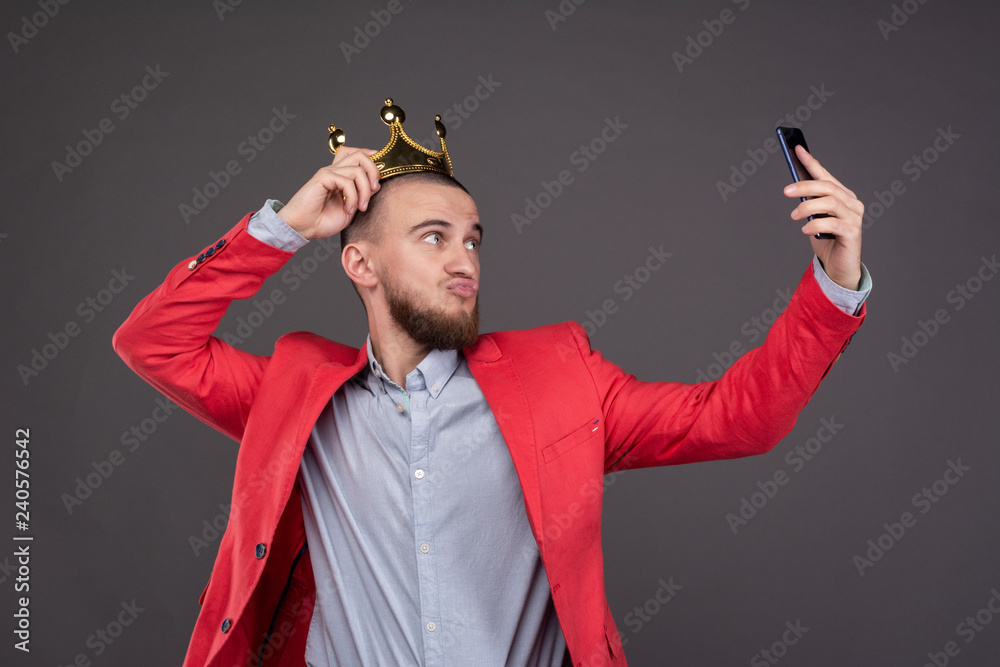 Young bearded handsome man in gold crown taking selfie looking at smartphone - obrazy, fototapety, plakaty 