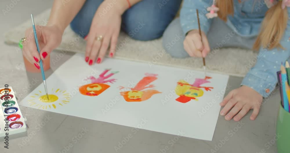 Close-up of hands of mother and little child with paintbrushes creating picture together on floor. Caring mother helping her cute girl developing creativity and artistic skills in paintings at home. Close-up of hands of mother and little child with paintbrushes creating picture together on floor. Caring mother helping her cute girl developing creativity and artistic skills in paintings at home.