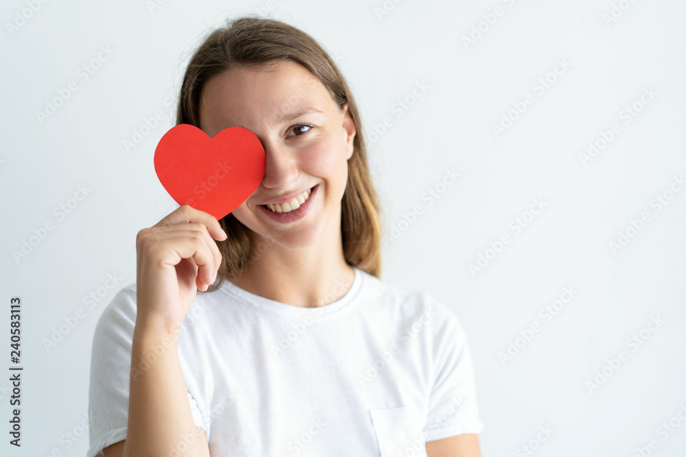 Playful young woman holding paper heart in front of her eye. Pretty girl looking at camera and smiling. Saint Valentines Day concept. Isolated front view on white background.