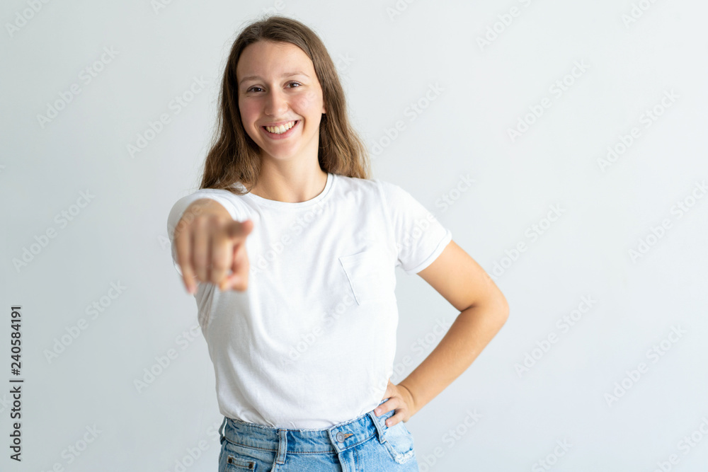 Obraz premium Smiling pretty young woman pointing at you and looking at camera. Confident girl holding hand on hip. Promotion concept. Isolated front view on white background.