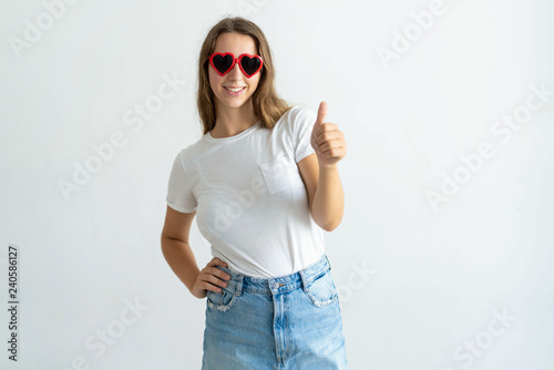Woman wearing heart shaped sunglasses and showing thumb up. Smiling pretty young lady looking at camera. Recommendation concept. Isolated front view on white background.