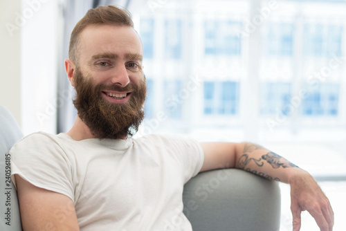Cheerful bearded hipster posing at home. Young man in casual resting on sofa and smiling at camera. Weekend at home concept