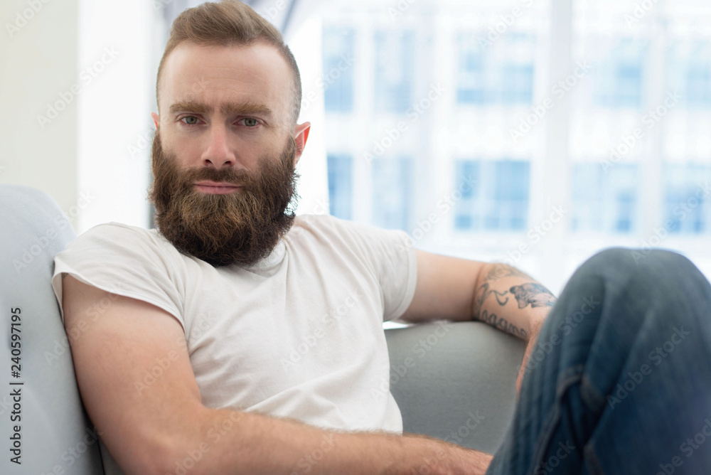 Pensive bearded guy posing at home. Young man in casual sitting on couch and staring at camera. Leisure at home concept