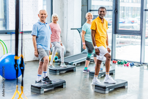 Fototapeta Naklejka Na Ścianę i Meble -  multiethnic senior athletes synchronous exercising on step platforms at gym