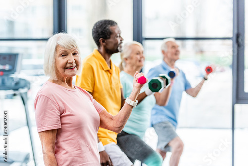 selective focus of smiling senior sportswoman and her friends exercising with dumbbells at sports hall