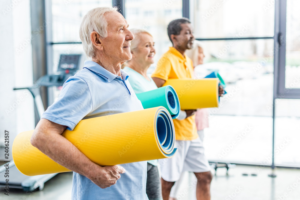 Fototapeta premium side view of multethnic senior athletes holding fitness mats at sports hall