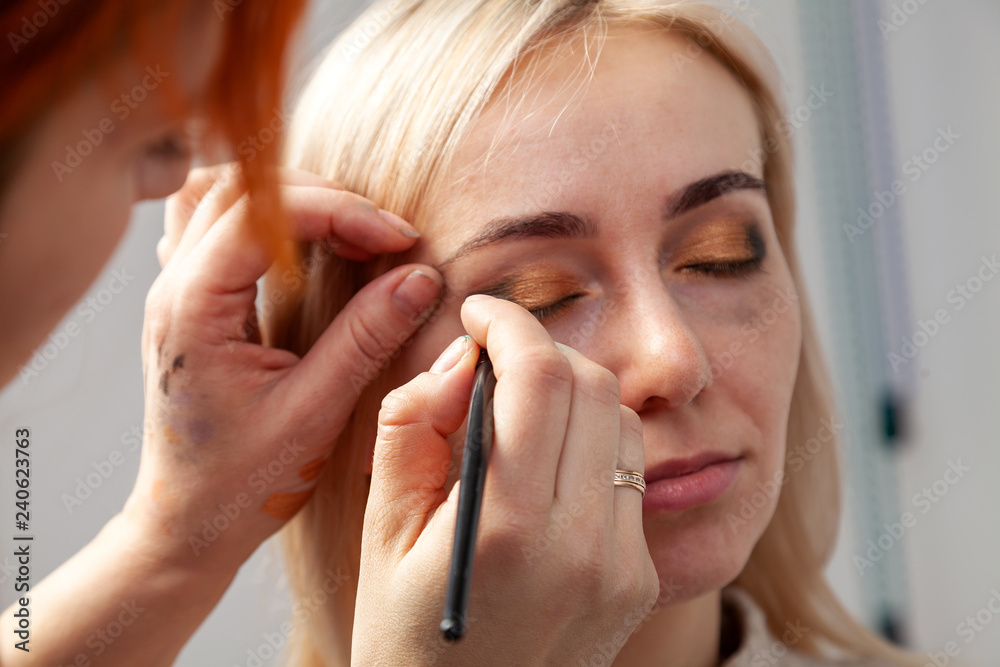 Close-up of applying make-up in the salon on the model in the oriental style, the make-up artist imposes golden brown shades and green in the corners of the century with a brush held in his hand.