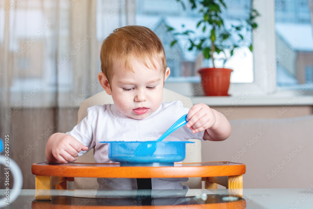 Little baby learning to eat with a spoon himself at the kids table in the kitchen. Healthy baby food Little baby learning to eat with a spoon himself at the kids table in the kitchen. Healthy baby food