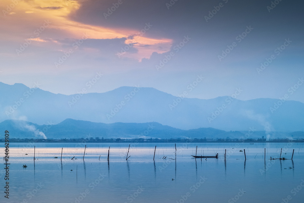 Fototapeta premium Lake view evening of a small fishing boat floating in the lake with mountain and cloudy sky background, sunset at Kwan Phayao, Phayao Province, northern of Thailand.