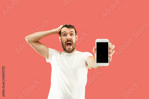 Fotografie Young handsome man showing smartphone screen over coral background with a surprise face