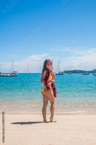 Back view of happy young woman with long hair in yellow bikini wearing funny merry christmas glasses and tinsel standing on tropical beach with yachts, blue water and sky on Phuket island,Thailand