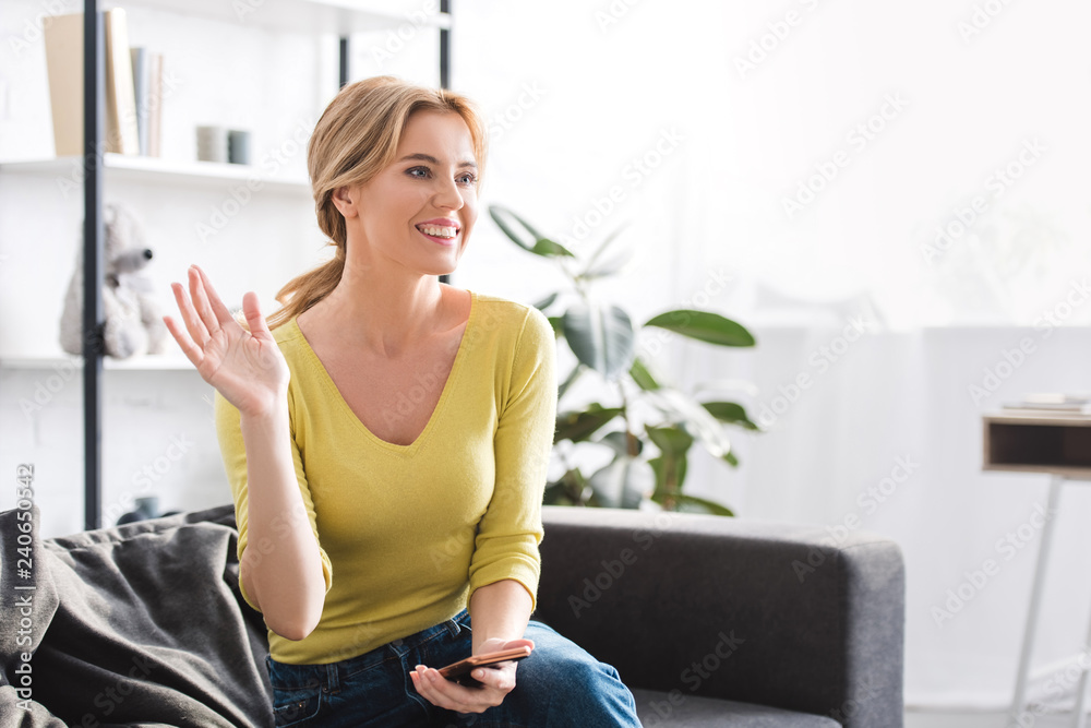 beautiful smiling woman holding smartphone and waving hand while sitting on couch at home beautiful smiling woman holding smartphone and waving hand while sitting on couch at home