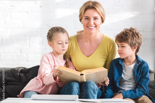 happy mother smiling at camera while reading book with adorable children at home