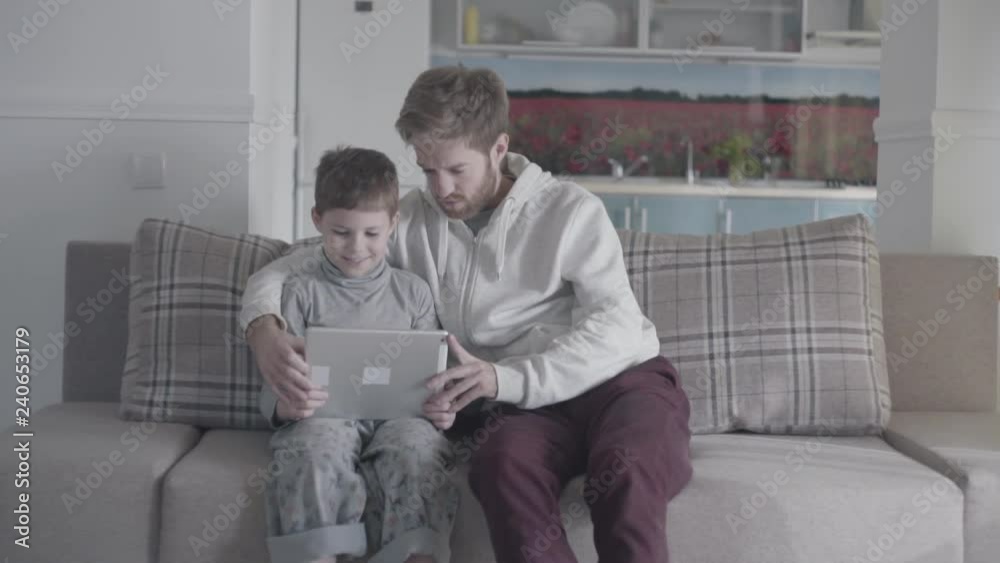 Bearded father and kid using digital tablet sitting on the couch in large living room. Dad teaches his son. Father-son relationship. Bearded father and kid using digital tablet sitting on the couch in large living room. Dad teaches his son. Father-son relationship.