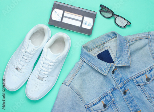 Retro style denim stylish jacket, video cassette, 3d glasses and white hipster sneakers on a mint-colored paper background. Minimalism, pop culture, top view.