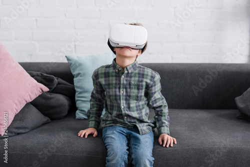 adorable child sitting on sofa and using virtual reality headset at home
