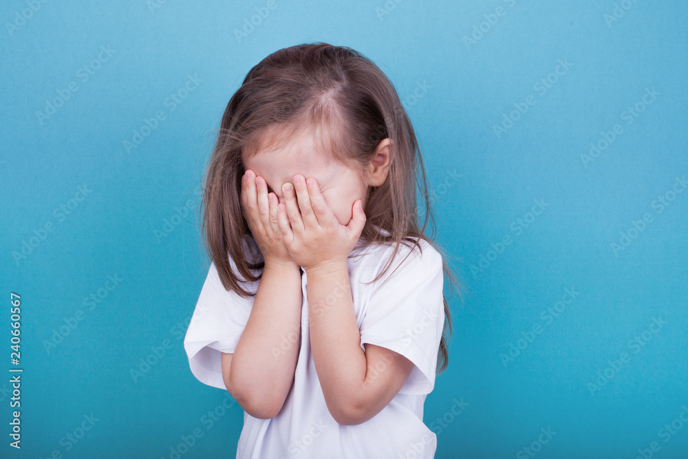 Little girl on a blue background crying covering her face with her hands - obrazy, fototapety, plakaty 