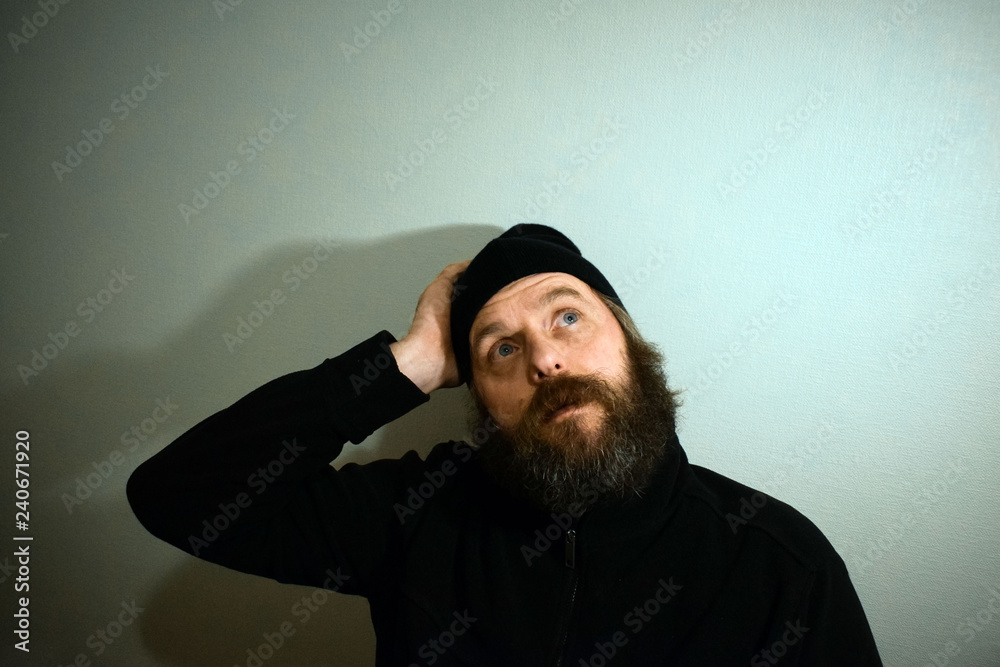 Bearded man 50 years old in black clothes and hat. A bearded man with blue eyes in a pensive pose is looking up. The concept of finding the answer to the question.