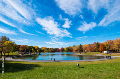 Beaver Lake on Mount Royal in Montreal, below a cloudy blue sky in autumn, with colorful maple trees and distant people around.