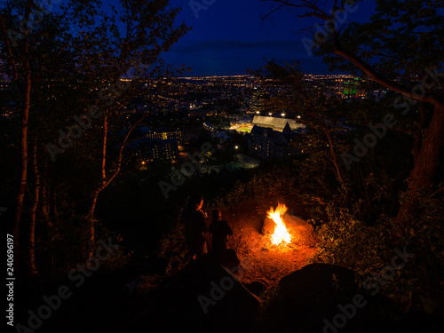 Two people looking at campfire overlooking city in the background at night.
